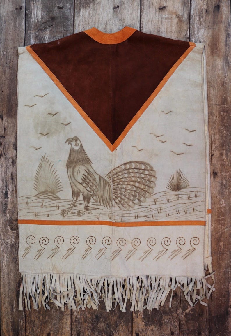 May include: A white leather poncho with a brown suede panel and orange trim. The poncho features a painted design of a bird with a long tail and a fringe border.