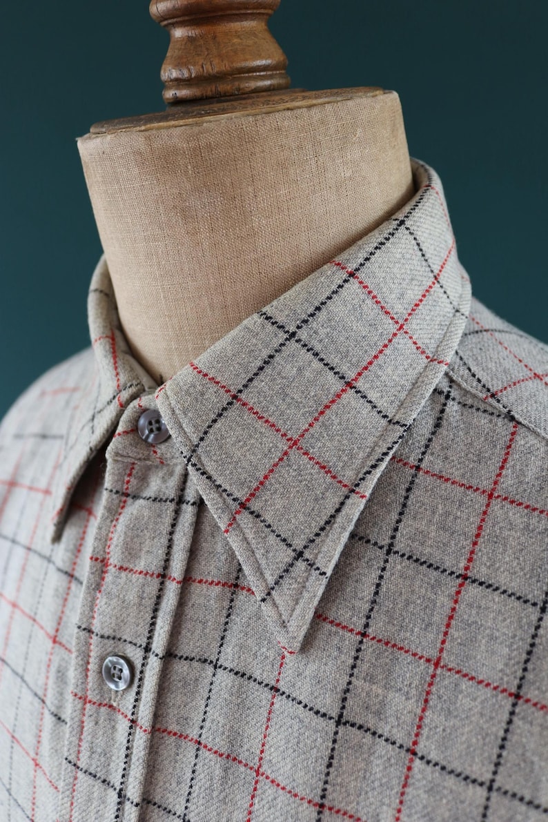 May include: A light gray button-down shirt with a plaid pattern of black and red lines. The shirt is displayed on a mannequin. The collar and button details are visible. The fabric appears to be a soft, woven material.