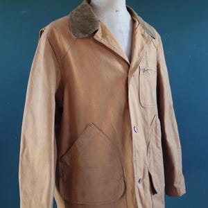 May include: A tan-colored vintage jacket with a corduroy collar, displayed on a mannequin. The jacket features large front pockets, a button closure, and a small chest pocket. The jacket's design suggests it is a workwear or outdoor garment.