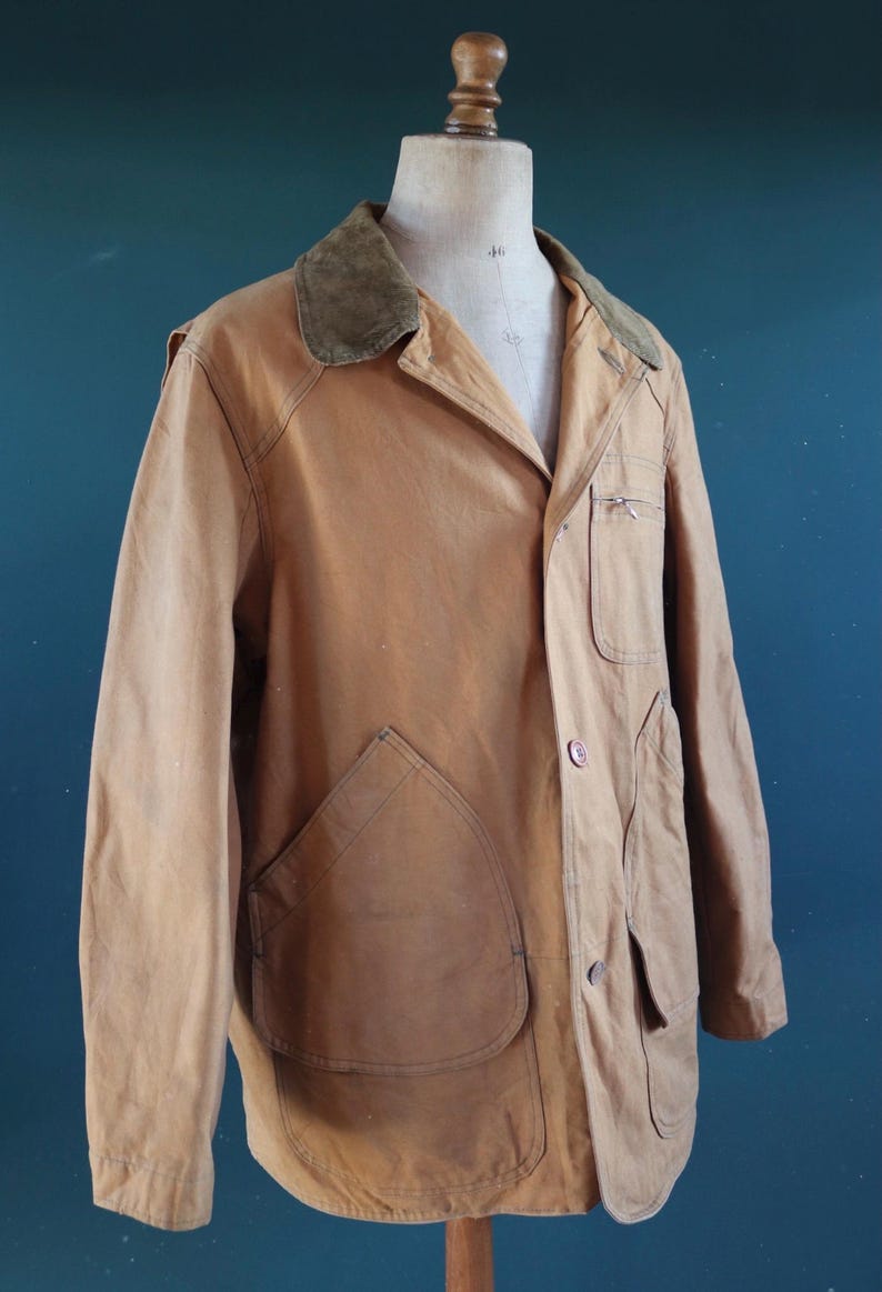 May include: A tan-colored vintage jacket with a corduroy collar, displayed on a mannequin. The jacket features large front pockets, a button closure, and a small chest pocket. The jacket's design suggests it is a workwear or outdoor garment.