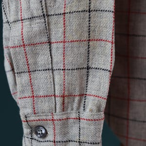 May include: Close-up of a gray button-down shirt sleeve. The fabric has a plaid pattern with red and black lines. A single gray button fastens the cuff. The shirt appears to be made of a textured material.