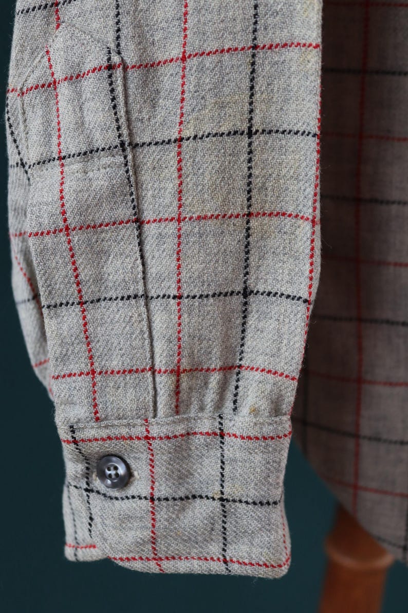 May include: Close-up of a gray button-down shirt sleeve. The fabric has a plaid pattern with red and black lines. A single gray button fastens the cuff. The shirt appears to be made of a textured material.