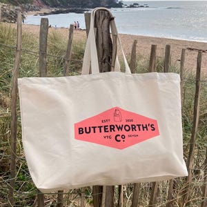 May include: A large, white canvas tote bag with a pink diamond-shaped logo that says "Butterworth's VTG Co. Devon" and "EST'D 2020". The bag is hanging on a wooden post on a beach.
