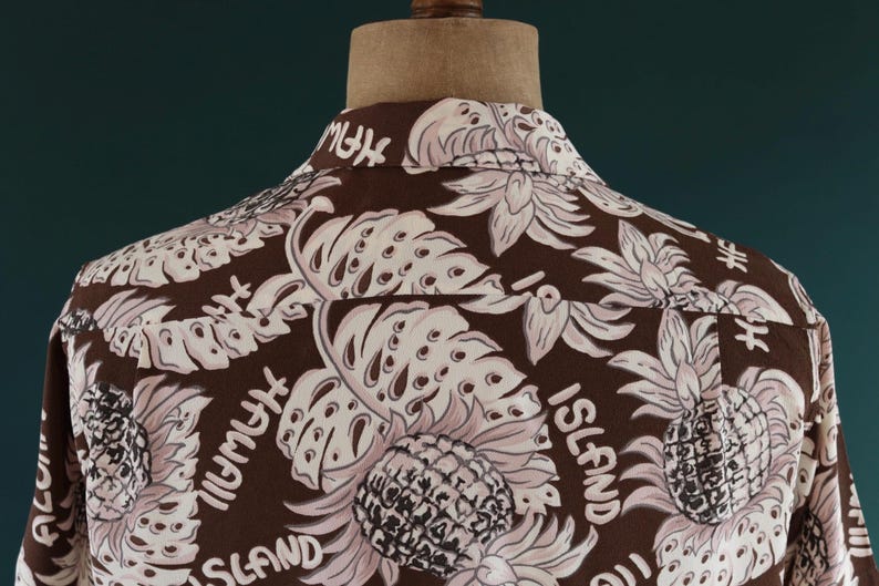 Peut inclure: Chemise boutonn&eacute;e marron &agrave; imprim&eacute; tropical avec des ananas, des feuilles et le texte "Island" et "High My" r&eacute;p&eacute;t&eacute; sur tout le motif.