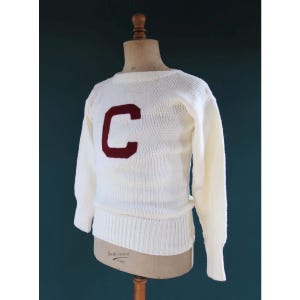 May include: Cream-colored knit sweater with a maroon "C" on the front. The sweater has long sleeves and a ribbed hem and cuffs. The garment is displayed on a mannequin against a dark teal background.