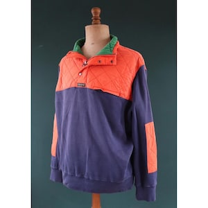 Peut inclure: Un sweat-shirt à encolure montante bicolore avec un empiècement et un coude orange matelassé, un corps bleu marine et un col vert. Le sweat-shirt a une encolure boutonnée et un petit écusson logo.