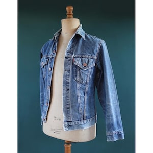May include: A vintage denim jacket in a medium wash blue, displayed on a mannequin. The jacket features a classic collar, button-up front, two chest pockets, and a red tag. The jacket is worn and faded, giving it a classic, timeless look.