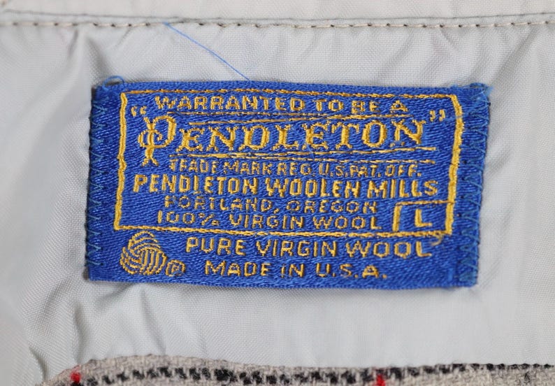 May include: A blue rectangular label with gold lettering and a gold border. The label reads "PENDLETON" and "100% VIRGIN WOOL" and "MADE IN U.S.A.". The label is sewn onto a light-colored fabric.