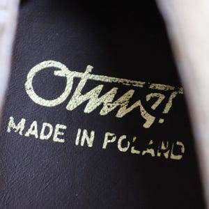 May include: Close-up of a dark brown leather surface with the gold text "Otus!" and "MADE IN POLAND" printed on it. The text is slightly worn, suggesting the item is vintage or well-used.