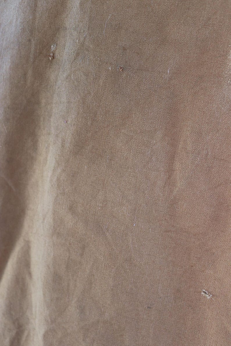 May include: Close-up of a textured, tan fabric with visible imperfections. The fabric has a slightly wrinkled appearance and a subtle sheen. Small, irregular marks are scattered across the surface.