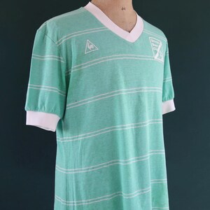 May include: A vintage, mint green t-shirt with white horizontal stripes. The shirt has a V-neck with a white collar and white cuffs. A small logo is visible on the chest. The shirt is displayed on a mannequin.