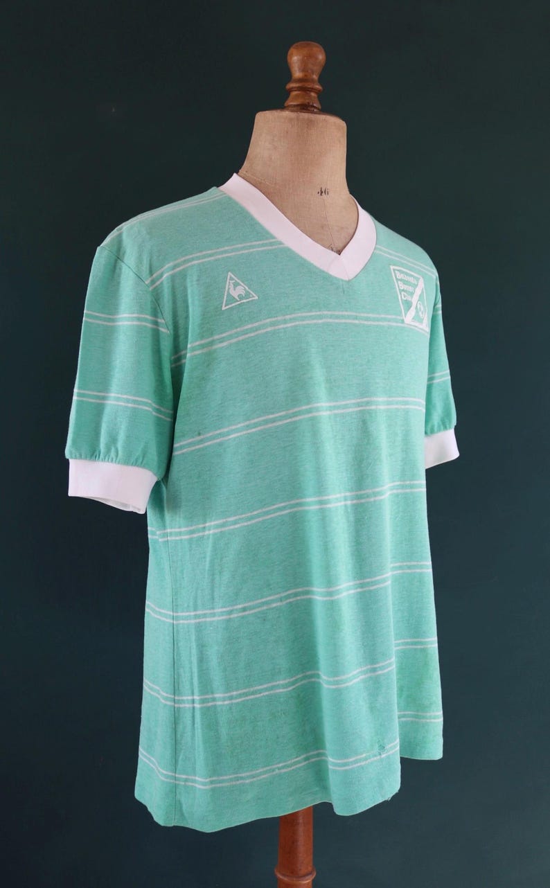 May include: A vintage, mint green t-shirt with white horizontal stripes. The shirt has a V-neck with a white collar and white cuffs. A small logo is visible on the chest. The shirt is displayed on a mannequin.