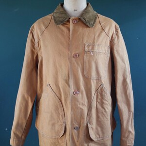 May include: A tan-colored jacket with a corduroy collar, button-up front, and multiple pockets. The jacket is displayed on a mannequin. The jacket has two large front pockets and two smaller pockets. The jacket is a classic style.