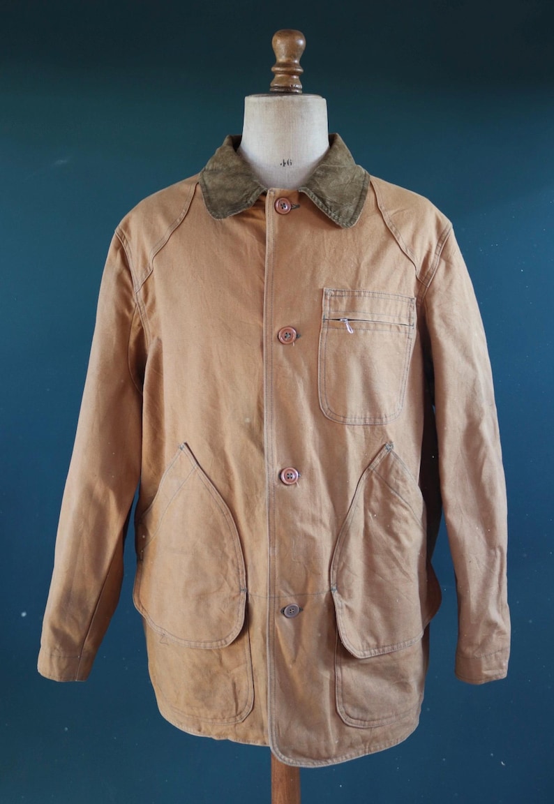 May include: A tan-colored jacket with a corduroy collar, button-up front, and multiple pockets. The jacket is displayed on a mannequin. The jacket has two large front pockets and two smaller pockets. The jacket is a classic style.