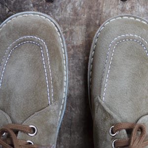 May include: A pair of suede shoes in a muted olive green hue. The shoes feature a rounded toe design with white stitching and brown laces. The soles are a light color, and the shoes are viewed from above.