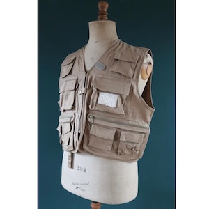 May include: A beige vest featuring numerous pockets and a zip fastening, displayed on a mannequin. The vest is constructed from a robust material and includes pockets of various sizes and shapes. Ideal for outdoor pursuits or practical use, it has a classic design.