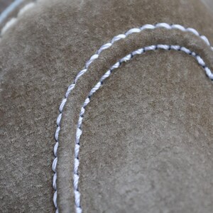 May include: Close-up of a section of a suede shoe in a muted olive green color. The shoe features white stitching along the edges and seams, creating a contrasting detail. The stitching is evenly spaced and adds a decorative element to the footwear.