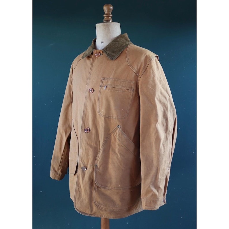 May include: A tan work jacket with a green corduroy collar, button closure, and multiple pockets. The jacket is displayed on a mannequin and has a classic, utilitarian design. The jacket is made of a durable, possibly waxed, material.