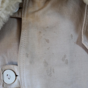 May include: Close-up of a tan jacket with a cream-colored, fluffy collar. The jacket has a button closure and visible staining on the fabric. The stitching is visible along the seams.