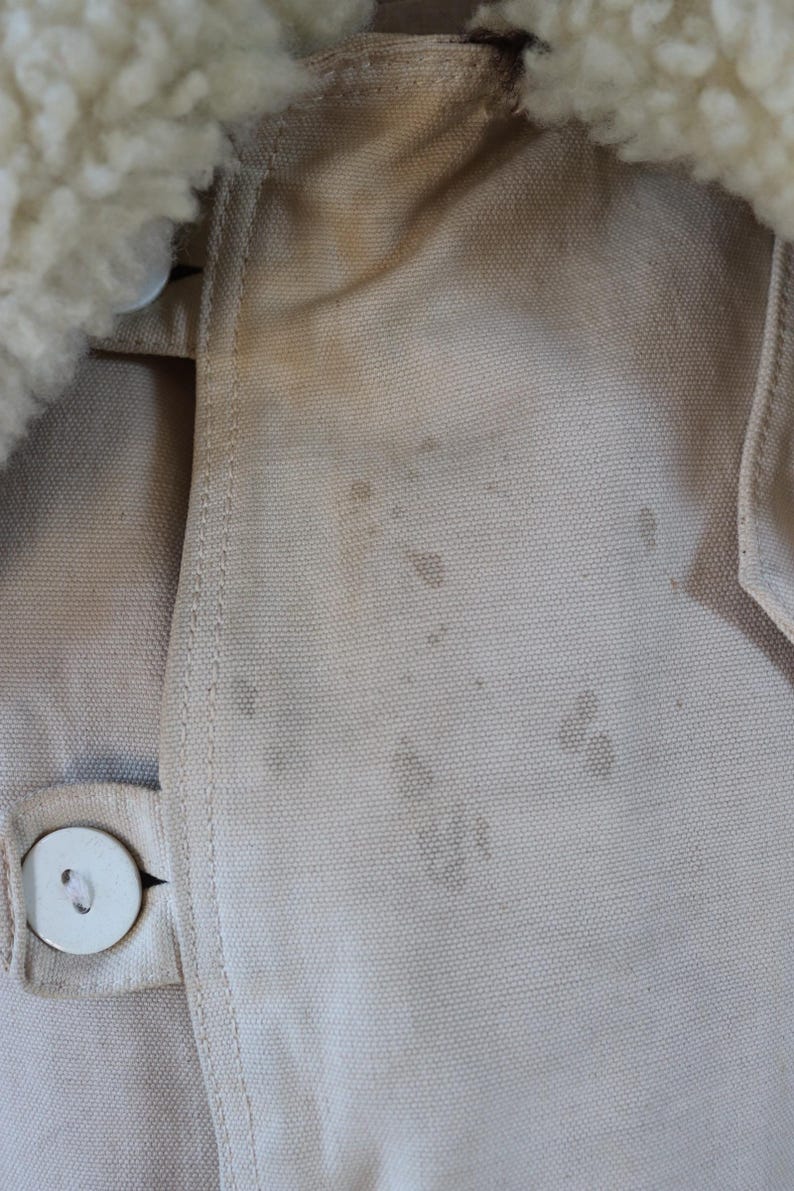 May include: Close-up of a tan jacket with a cream-colored, fluffy collar. The jacket has a button closure and visible staining on the fabric. The stitching is visible along the seams.