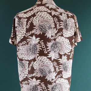 Peut inclure: Une chemise &agrave; manches courtes marron avec un motif de feuilles et d'ananas blancs et roses. La chemise porte le texte "Aloha" et "Hawaii Island" r&eacute;p&eacute;t&eacute; sur tout le motif.