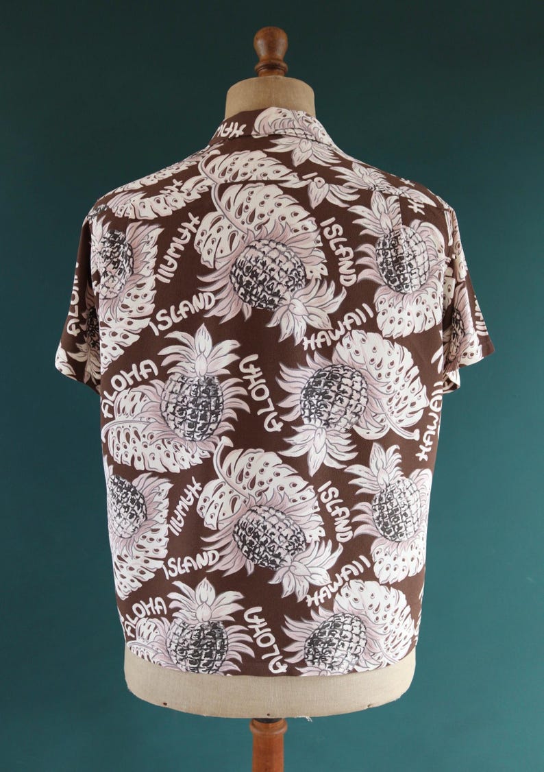 Peut inclure: Une chemise &agrave; manches courtes marron avec un motif de feuilles et d'ananas blancs et roses. La chemise porte le texte "Aloha" et "Hawaii Island" r&eacute;p&eacute;t&eacute; sur tout le motif.