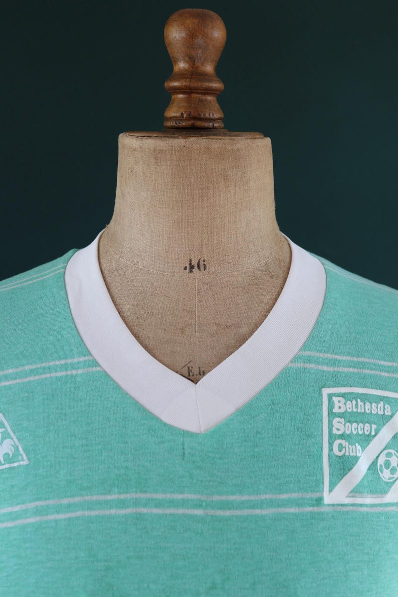 May include: A vintage-style teal soccer jersey with a white V-neck collar and horizontal stripes. The jersey features the "Bethesda Soccer Club" logo on the right chest and a small emblem on the left. The jersey is displayed on a mannequin.