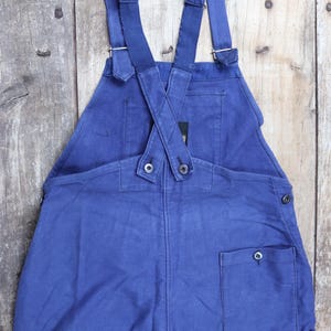 May include: Blue denim dungarees with adjustable straps and a single back pocket. The straps are crossed at the back and fastened with buttons.