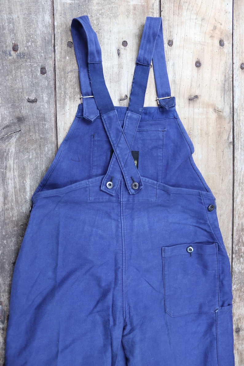 May include: Blue denim dungarees with adjustable straps and a single back pocket. The straps are crossed at the back and fastened with buttons.