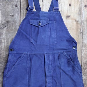 May include: A pair of blue denim dungarees with a large front pocket and two smaller side pockets. The dungarees have adjustable straps with metal buckles.