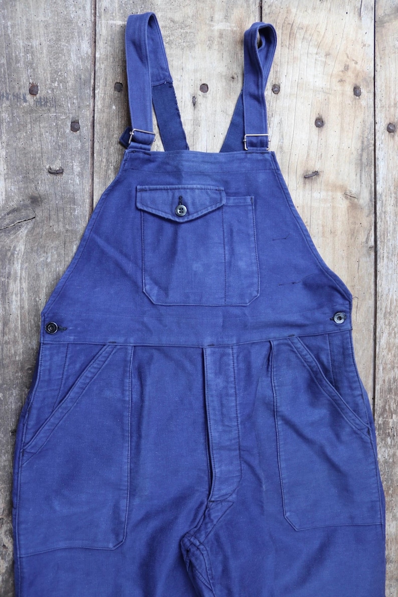 May include: A pair of blue denim dungarees with a large front pocket and two smaller side pockets. The dungarees have adjustable straps with metal buckles.