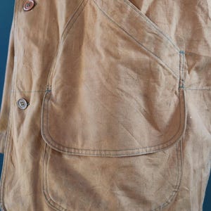 May include: Close-up of a tan work jacket with large patch pockets and button closures. The jacket has visible stitching in a contrasting blue thread. The fabric appears to be a durable, possibly canvas material, with a slightly worn appearance.