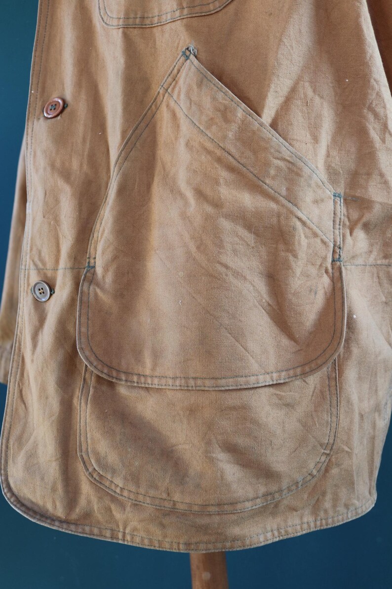 May include: Close-up of a tan work jacket with large patch pockets and button closures. The jacket has visible stitching in a contrasting blue thread. The fabric appears to be a durable, possibly canvas material, with a slightly worn appearance.