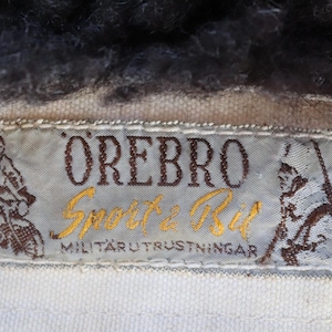 May include: Close-up of a label with the text "&Ouml;REBRO Sport & Bil MILIT&Auml;RUTRUSTNINGAR". The label is light blue with brown and gold text and illustrations of figures. The label is sewn onto a white fabric, with dark fur at the top.