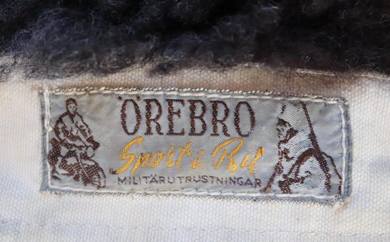 May include: Close-up of a label with the text "&Ouml;REBRO Sport & Bil MILIT&Auml;RUTRUSTNINGAR". The label is light blue with brown and gold text and illustrations of figures. The label is sewn onto a white fabric, with dark fur at the top.