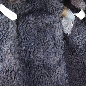 May include: Close-up of the inside of a sheepskin coat. The coat's interior features dark gray, curly wool with patches of lighter gray and cream-colored wool. White fabric strips are visible.