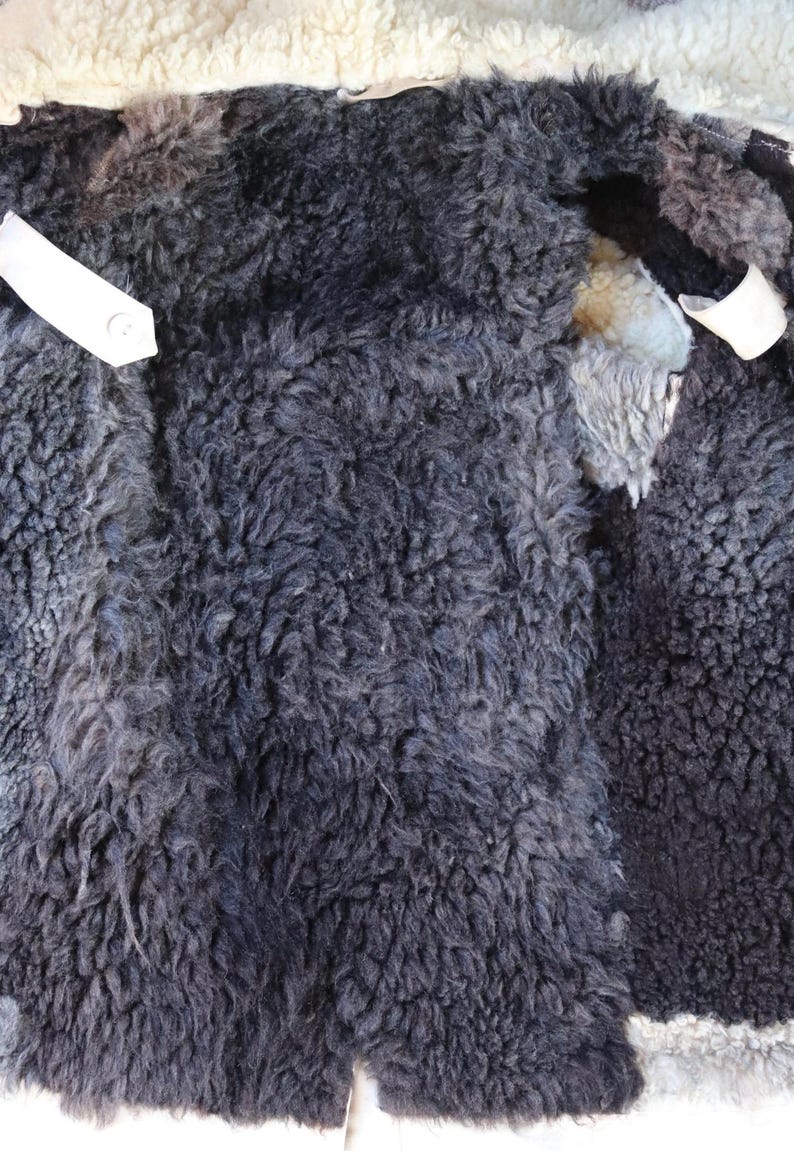 May include: Close-up of the inside of a sheepskin coat. The coat's interior features dark gray, curly wool with patches of lighter gray and cream-colored wool. White fabric strips are visible.