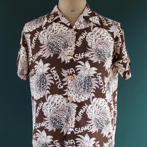 Peut inclure: Une chemise &agrave; manches courtes marron avec un motif d'ananas et de feuilles blanc et rose. La chemise porte les mots "Aloha", "Hawaii" et "Island" imprim&eacute;s dessus.