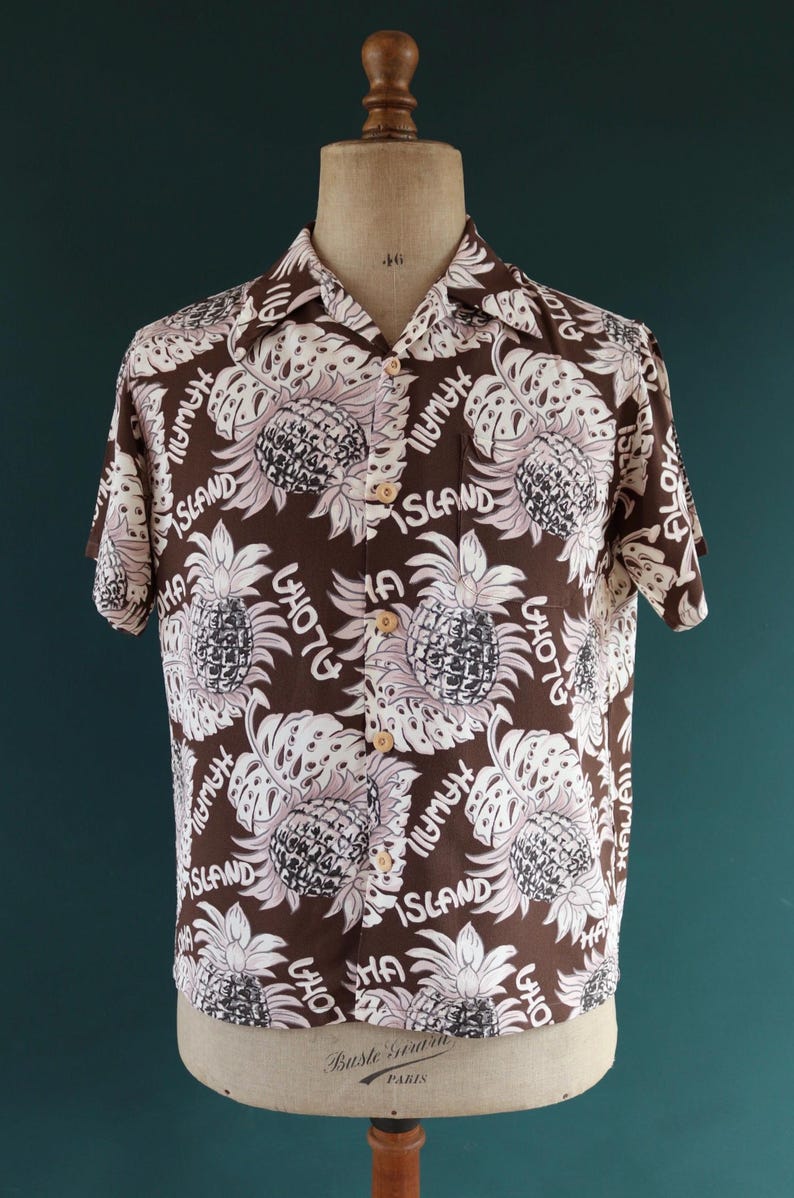 Peut inclure: Une chemise &agrave; manches courtes marron avec un motif d'ananas et de feuilles blanc et rose. La chemise porte les mots "Aloha", "Hawaii" et "Island" imprim&eacute;s dessus.