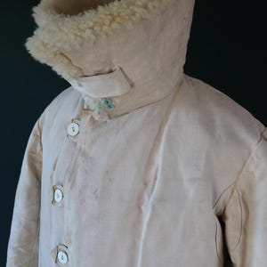 May include: Cream-colored vintage coat with a high collar lined with fluffy, off-white material. The coat features a button-up front with large, round buttons and a small tab detail on the collar. The coat appears to be made of a heavy, durable fabric.