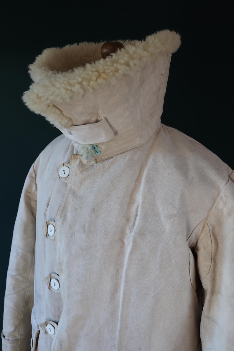 May include: Cream-colored vintage coat with a high collar lined with fluffy, off-white material. The coat features a button-up front with large, round buttons and a small tab detail on the collar. The coat appears to be made of a heavy, durable fabric.