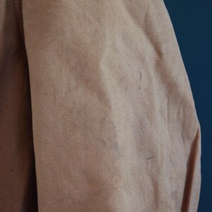 May include: Close-up of a tan fabric, possibly a jacket or coat, with visible texture and slight discoloration. The fabric appears to be a sturdy material, with a subtle sheen. The image focuses on the shoulder and sleeve area.