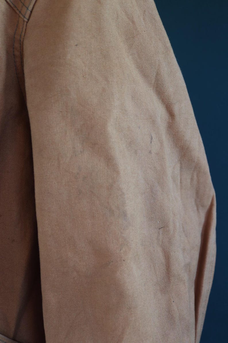 May include: Close-up of a tan fabric, possibly a jacket or coat, with visible texture and slight discoloration. The fabric appears to be a sturdy material, with a subtle sheen. The image focuses on the shoulder and sleeve area.