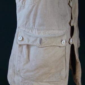 May include: Cream-colored, vintage-style coat with a button-up front and a large pocket. The coat features several white buttons and a tailored design. The fabric appears to be a sturdy, possibly linen or cotton blend, with visible wear.