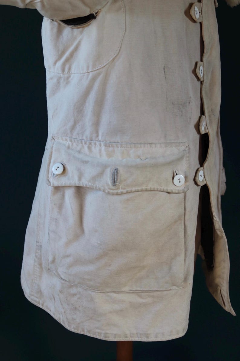 May include: Cream-colored, vintage-style coat with a button-up front and a large pocket. The coat features several white buttons and a tailored design. The fabric appears to be a sturdy, possibly linen or cotton blend, with visible wear.