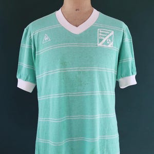 May include: A vintage teal soccer jersey with horizontal white stripes. The jersey has a V-neck collar and short sleeves with white cuffs. The text "Bethesda Soccer Club" is printed on the right chest.