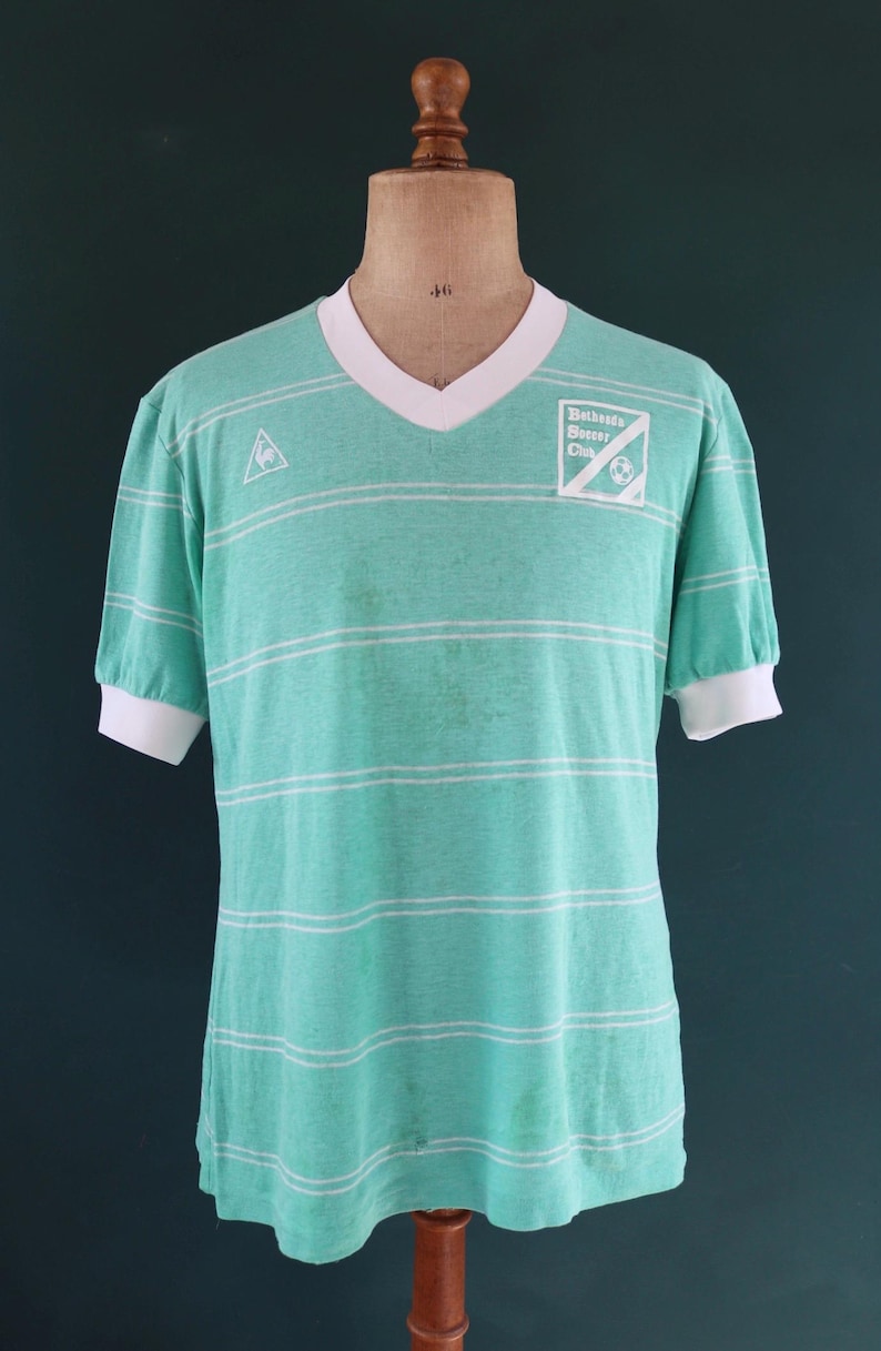 May include: A vintage teal soccer jersey with horizontal white stripes. The jersey has a V-neck collar and short sleeves with white cuffs. The text "Bethesda Soccer Club" is printed on the right chest.