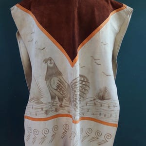 May include: A white leather vest with a brown suede panel and orange trim. The vest features a painted design of a bird with a long tail and a decorative border of swirls. The vest has fringe along the bottom.