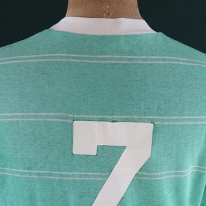 May include: A green and white striped t-shirt with the number 7 on the back. The shirt has a white collar and sleeve cuffs. The fabric appears to be a soft knit material.