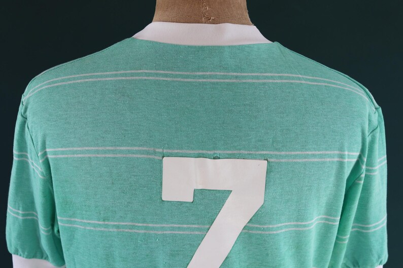 May include: A green and white striped t-shirt with the number 7 on the back. The shirt has a white collar and sleeve cuffs. The fabric appears to be a soft knit material.
